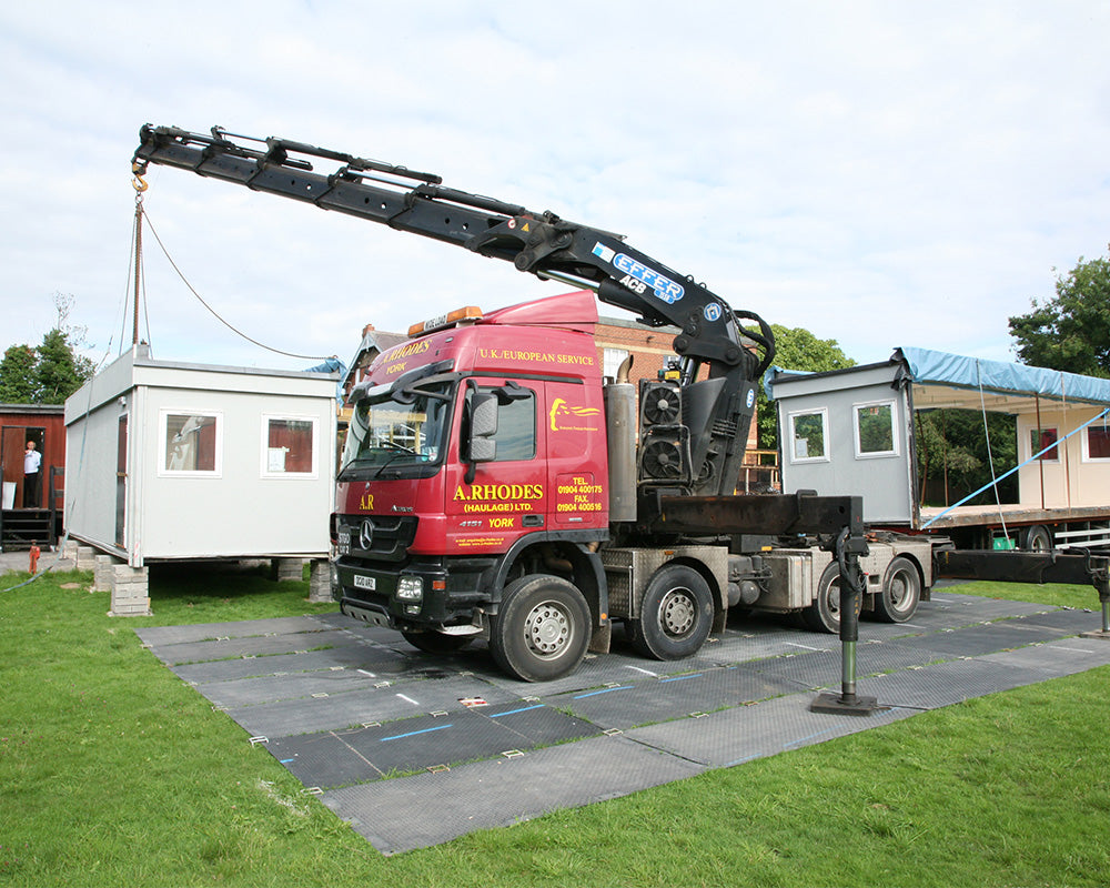 Truck with crane lifting a small building on a grassy area with buildings in the background.