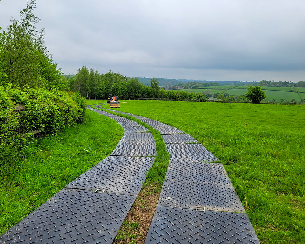 Winding path made of ground protection trackway in a green field with trees and mountains in the background.