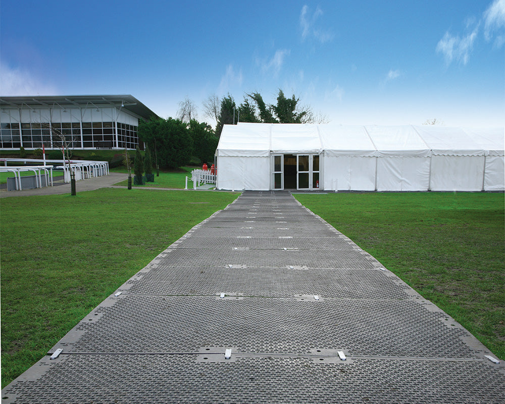 Plastic walkway leading to a large white tent on a grassy area with a building in the background.